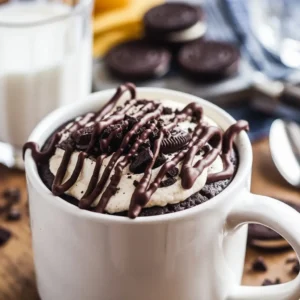 A white ceramic mug filled with a rich, chocolatey dessert, topped with a swirl of whipped cream and sprinkled with Oreo crumbs. The mug sits on a warm, inviting kitchen table with soft lighting. A spoon rests beside it, ready for a bite, while faint steam rises, indicating it's freshly made.