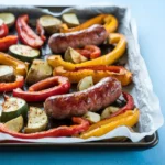 A sheet pan of golden-brown sausages and caramelized vegetables—bell peppers, zucchini, cherry tomatoes, and red onions—glistening with olive oil and topped with fresh rosemary and thyme. Set on a rustic wooden table with a cozy kitchen backdrop.