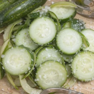 A close-up of thinly sliced fresh cucumbers sprinkled with salt on a wooden cutting board. The cucumbers glisten as they release moisture, with a small timer in the background indicating a 10-minute wait. A clean kitchen towel is nearby, ready for patting them dry.
